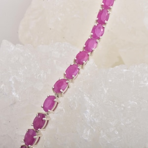 Pode incluir: Uma pulseira de prata com uma série de pedras preciosas rosa vibrantes de corte oval. As pedras são engastadas individualmente em engastes de prata, criando uma linha contínua de cor. A pulseira é exibida sobre um fundo branco texturizado.