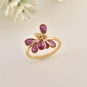 Puede incluir: Un anillo de oro con un grupo de piedras preciosas rojas en forma de lágrima. Las piedras están dispuestas en un diseño floral o abstracto, con un grupo central y pétalos circundantes. El anillo tiene una banda de oro pulido.