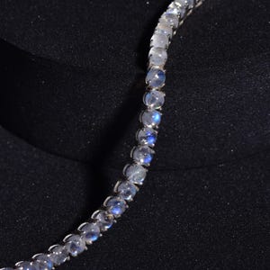Pode incluir: Um colar delicado com contas redondas de pedra da lua iridescentes. As contas são fixadas em metal prateado, criando um efeito brilhante. O colar é exibido em um fundo escuro.