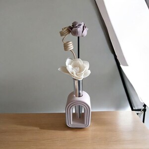Peut inclure: Un diffuseur de roseaux décoratif avec trois fleurs artificielles. Les fleurs sont une rose violet clair, une spirale crème et un magnolia blanc. Le diffuseur est dans un vase en céramique rose clair en forme d'arche avec un tube argenté.