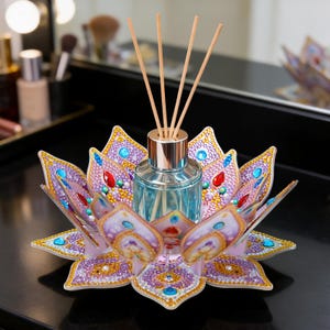 May include: A decorative lotus flower-shaped diffuser holder with a glass bottle and wooden reeds. The holder is adorned with colorful rhinestones in shades of purple, blue, and gold. The bottle contains a light blue liquid.