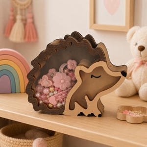 May include: A wooden hedgehog-shaped piggy bank with a clear acrylic front panel. The interior is filled with various pink and white decorative beads, flower-shaped hair clips, and heart-shaped accessories. The hedgehog has a dark brown frame.