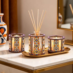 May include: Decorative set featuring a glass diffuser with reeds and two candle holders. The holders and diffuser are adorned with a colorful geometric pattern in blue, yellow, and orange. The set sits on a gold tray, creating a warm, inviting ambiance.