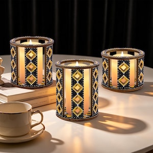 May include: Three decorative candle holders with lit candles. The holders are cylindrical with a wooden frame and are decorated with black, gold, and clear beads in a diamond pattern. A teacup and saucer are in the foreground.