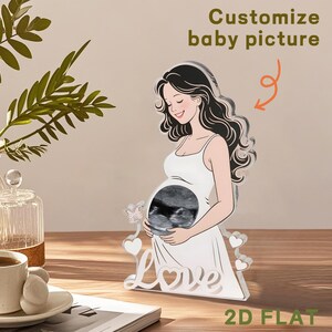 May include: A clear acrylic standee featuring an illustration of a pregnant person in a white dress with a circular cutout over the belly displaying a black and white ultrasound image. The base includes the word Love and small heart and butterfly accents.