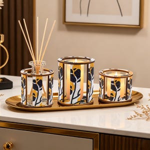 May include: A decorative set of three items on a gold tray. The set includes a reed diffuser with wooden sticks, and two candle holders. The candle holders and diffuser are decorated with black, yellow, and white floral patterns.