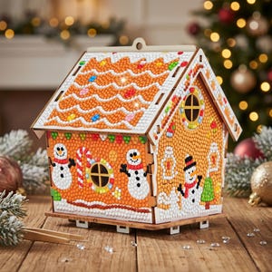 May include: A decorative gingerbread house craft kit featuring a diamond-painted surface with snowman, candy cane, and tree motifs. The structure has a light brown base with white icing details, a peaked roof, and a circular window on the front.