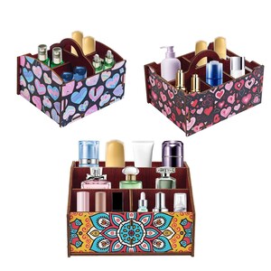 Op de afbeelding: Drie houten cosmetica-organizers. De bovenste twee zijn zwart met hartpatronen en de onderste is bruin met een bloemmotief. Elke organizer heeft meerdere compartimenten en een handvat. De organizers zijn gevuld met diverse cosmetische producten.