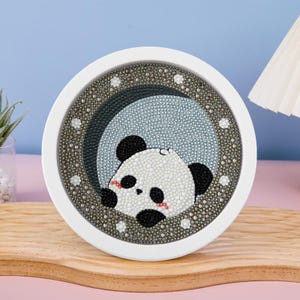 May include: A round, white-framed artwork features a panda bear design. The panda is depicted with black and white details, accented with red cheeks. The background is a light blue hue, and the frame is embellished with small, sparkling accents. The artwork is displayed on a wooden surface.