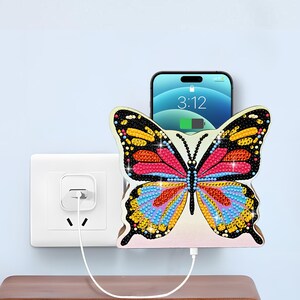 May include: A wall-mounted smartphone charging holder shaped like a butterfly, featuring a colorful diamond-art style mosaic pattern with black, pink, yellow, and blue accents. The holder is plugged into a white wall outlet with a charging cable.