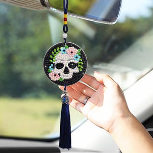May include: A round car charm featuring a skull design with floral accents in pink, blue, and green. The charm is embellished with sparkling rhinestones and has a navy blue tassel. It hangs from a rearview mirror.