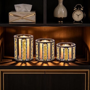 May include: Three decorative candle holders with lit candles. The holders have a cylindrical shape with a geometric pattern in gold, black, and white. The candles are placed on a dark wooden shelf with other decorative items.