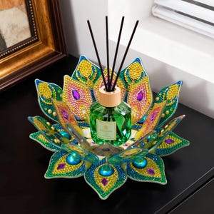 May include: A decorative, jewel-encrusted lotus flower-shaped tray holds a green glass diffuser bottle with black reeds. The tray features a peacock feather design with blue, yellow, and purple accents. The diffuser has a wooden cap.