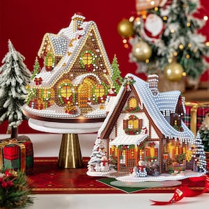 Puede incluir: Dos casas navideñas decorativas con adornos brillantes. Una casa está sobre un soporte dorado, la otra sobre una base. Ambas tienen tejados cubiertos de nieve, ventanas iluminadas y detalles festivos como coronas y árboles en miniatura. Las casas están sobre un mantel rojo.