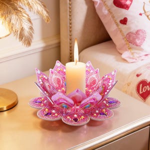 Op de afbeelding: Een brandende kaars staat in een roze lotusbloemvormige kandelaar. De kandelaar is versierd met strass-steentjes en staat op een goudkleurig nachtkastje. De achtergrond bevat een bed met roze beddengoed met harten.