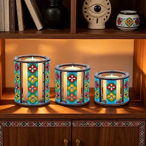 May include: Set of three decorative candle holders with colorful floral patterns. The candle holders are cylindrical with a wooden frame and glass panels. Each holder has a lit candle inside, creating a warm glow. The design features red, yellow, and blue floral motifs.