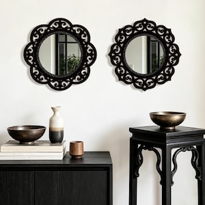 May include: Two black decorative mirrors with ornate, floral frames on a white wall. Below, a black cabinet displays a bronze bowl, books, a vase, and a wooden cup. A black side table holds a bronze bowl.