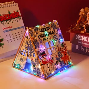 May include: A decorative Christmas house with the words "MERRY CHRISTMAS" on the roof. The house is adorned with colorful lights, snowflakes, and a snowman. A calendar showing the month of December is in the background.