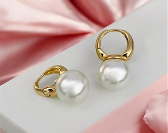 Puede incluir: Un par de pendientes de aro dorados, cada uno con una perla blanca redonda y grande. Los pendientes se exhiben sobre una superficie blanca contra un fondo de tela rosa. Los pendientes tienen un diseño clásico y elegante.