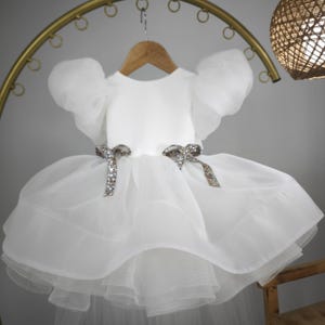 May include: A white dress with a layered skirt and puffy sleeves. The dress features two decorative bows with sparkling embellishments at the waist. The dress is hanging on a wooden hanger.
