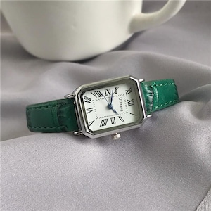 Puede incluir: Un reloj de pulsera rectangular plateado con esfera blanca y números romanos negros. El reloj tiene una correa de cuero verde. La esfera del reloj tiene la palabra "Ulzzang" impresa. El reloj está sobre una superficie gris.