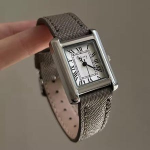 May include: A silver rectangular wristwatch with a grey leather strap. The watch face is white with black Roman numerals and the brand name "Goey". The strap is textured. The watch is held by a hand.