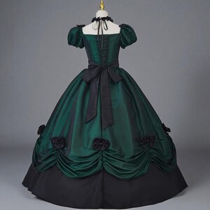 May include: A green and black Victorian-style gown with a fitted bodice, short puffed sleeves, and a full skirt. The dress features a black bow at the waist and decorative black rosettes on the skirt. The bodice has a lace-up detail.