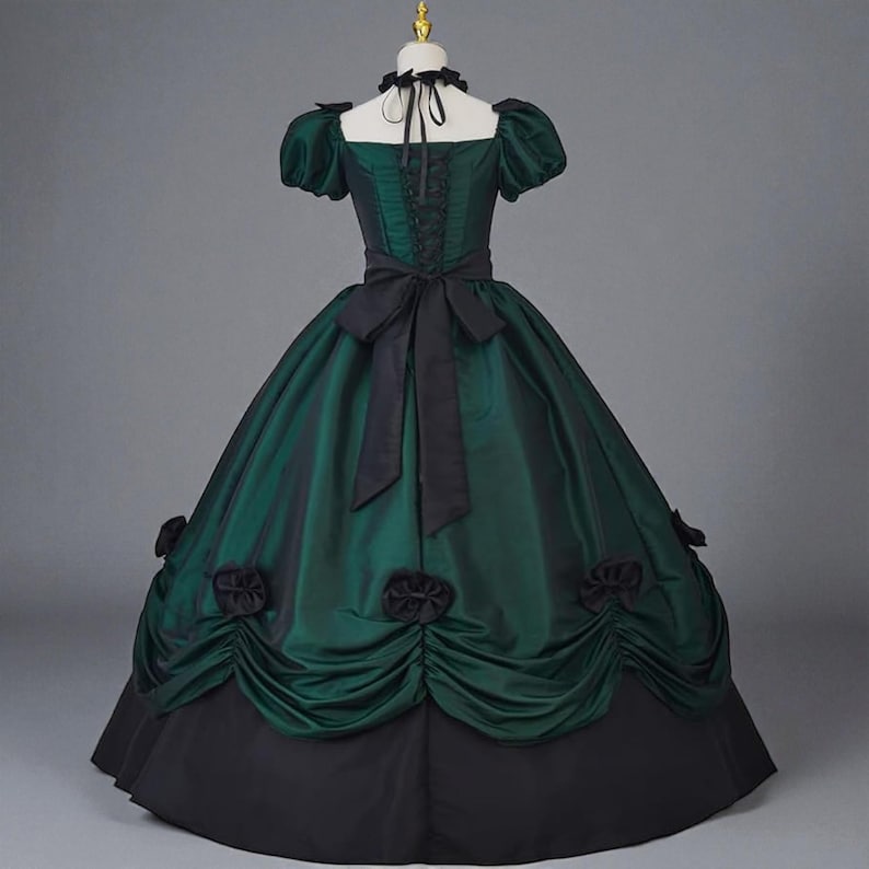 May include: A green and black Victorian-style gown with a fitted bodice, short puffed sleeves, and a full skirt. The dress features a black bow at the waist and decorative black rosettes on the skirt. The bodice has a lace-up detail.