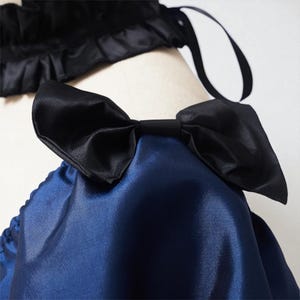 May include: Close-up of a navy blue satin garment with a large black bow. The bow is centered and prominent, with a smooth, shiny texture. A black satin trim with a ruffled edge is visible in the background.