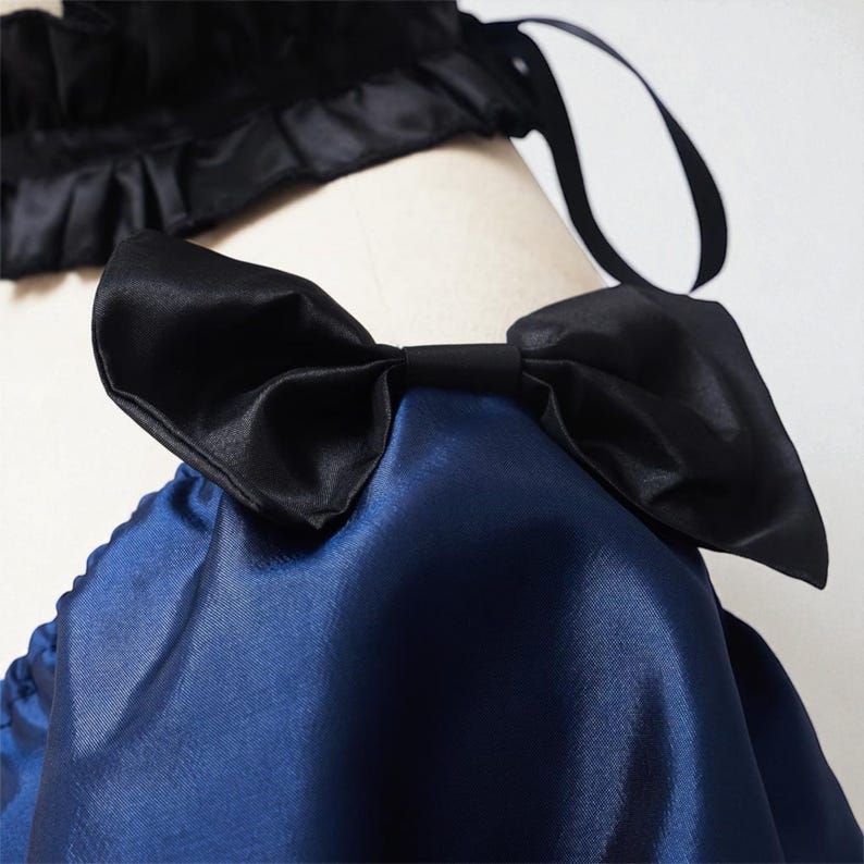 May include: Close-up of a navy blue satin garment with a large black bow. The bow is centered and prominent, with a smooth, shiny texture. A black satin trim with a ruffled edge is visible in the background.