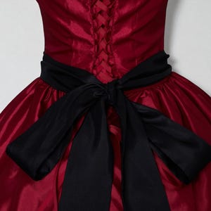 May include: A formal red dress with a black satin bow. The dress features a corset back and a full skirt. The bow is tied at the waist, adding a contrasting accent to the dress's design. The fabric has a slight sheen.