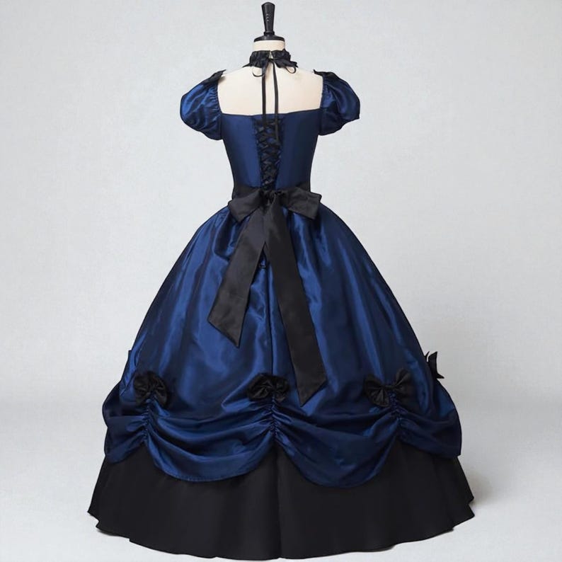 May include: A navy blue ball gown with a black skirt and accents. The dress features a square neckline, short puffed sleeves, and a black bow at the waist. Black rosettes adorn the skirt.