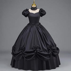 May include: A black Victorian-style gown with a high ruffled collar and short, puffed sleeves. The full skirt features decorative bows and gathered fabric, creating a classic silhouette. The dress is displayed on a mannequin.