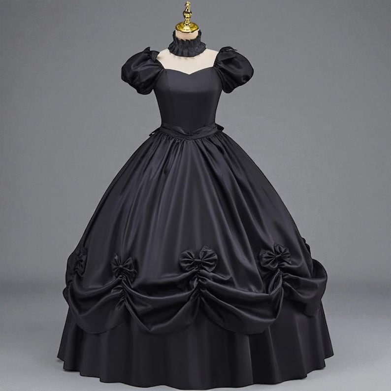 May include: A black Victorian-style gown with a high ruffled collar and short, puffed sleeves. The full skirt features decorative bows and gathered fabric, creating a classic silhouette. The dress is displayed on a mannequin.