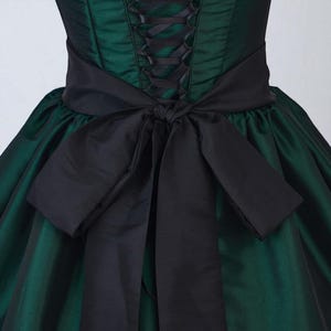 May include: A dark green dress with a black corset-style bodice and a large black bow at the waist. The dress has a full skirt and a satin-like sheen, creating a formal and elegant appearance. The bodice features black lacing.