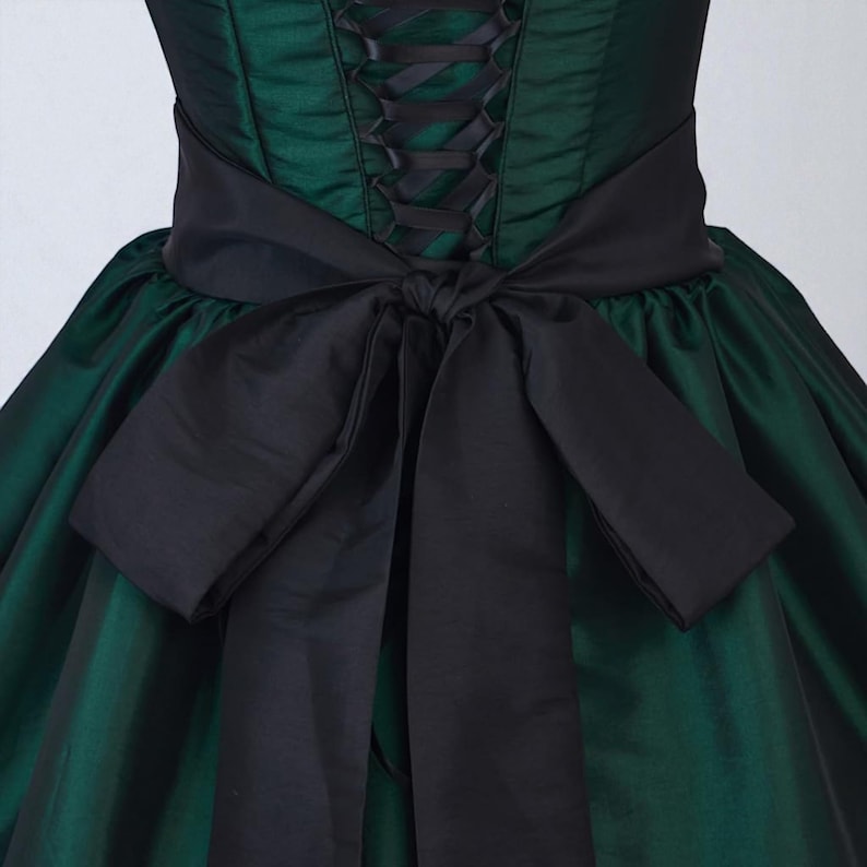 May include: A dark green dress with a black corset-style bodice and a large black bow at the waist. The dress has a full skirt and a satin-like sheen, creating a formal and elegant appearance. The bodice features black lacing.
