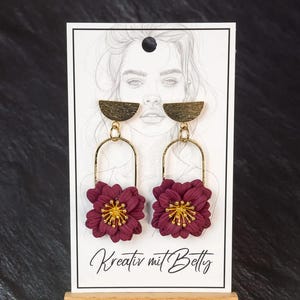 Könnte beinhalten: Burgunderfarbene Blumenohrringe mit goldenen Akzenten. Die Ohrringe haben einen goldenen Halbkreis-Aufsatz, einen goldenen Bogen und eine burgunderfarbene Blume mit goldenem Zentrum. Sie werden auf einer weißen Karte mit dem Text "Kreativ mit Betty" präsentiert.