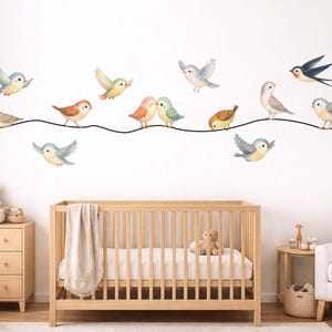 May include: A nursery scene featuring a light wood crib, dresser, and a white armchair. The wall is decorated with watercolor-style bird decals in various colors, perched on a black line. A teddy bear and other decor complete the room.