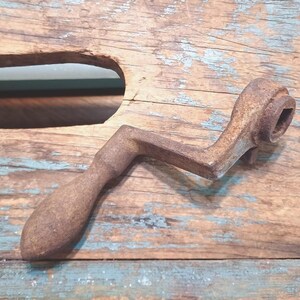 Peut inclure: Une manivelle en métal rouillé et vintage avec une poignée en bois, posée sur une surface en bois vieilli. La manivelle a une douille carrée à une extrémité et un design incurvé. La surface en bois a un aspect vieilli avec des tons bleus et bruns.