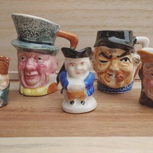 May include: Five ceramic character jugs with detailed faces. The jugs feature a variety of designs, including a man in a top hat and a figure in a blue coat. The jugs are displayed on a wooden surface.