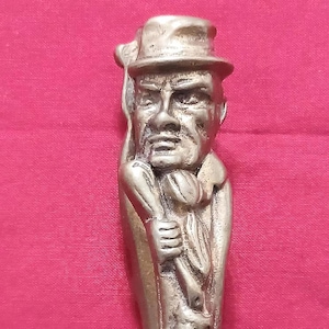 May include: A metallic silver-toned decorative object featuring a sculpted relief of a figure wearing a hat and a coat, holding a long, slender item in one hand. The detailed casting shows facial features, clothing textures, and a smooth finish.