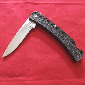 May include: A folding knife with a silver blade and a black textured handle. The blade is partially open, showing the sharp edge. The knife is set against a red background. The brand "BUCK" is visible on the blade.