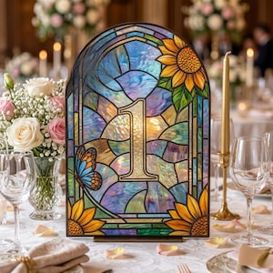 Puede incluir: Un letrero de mesa de vitral en forma de arco con el número 1 en el centro, decorado con girasoles amarillos y una mariposa azul. Los paneles de vidrio muestran tonos iridiscentes azules, morados y verdes con un marco de metal oscuro.