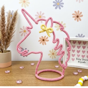 Puede incluir: Una silueta de cabeza de unicornio tejida en rosa montada sobre una base circular, con un lazo amarillo tejido en la frente y una cuenta transparente cerca del hocico, hecha de hilo en una forma estructurada y continua.