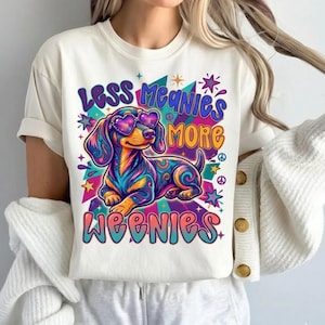 Puede incluir: Una camiseta de color crema con un gráfico colorido de un perro salchicha con gafas de sol en forma de corazón. El diseño incluye patrones de remolinos en azul, naranja y morado, con el texto "LESS MEANIES MORE WEENIES" en letras negritas.