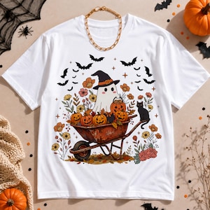 Puede incluir: Una camiseta blanca de manga corta con un estampado gráfico de un fantasma con sombrero de bruja, sentado en una carretilla oxidada llena de calabazas de Halloween y flores de otoño, junto a un gato negro y murciélagos volando.