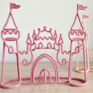 Puede incluir: Una escultura de alambre tejido en color rosa con forma de castillo, que cuenta con dos torres altas rematadas por pequeñas banderas. La estructura presenta una puerta central arqueada, torretas decorativas y un recubrimiento de lana tejida.