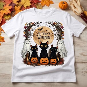 Puede incluir: Una camiseta blanca con un estampado gráfico de tres gatos negros sentados con calabazas de Halloween, dos fantasmas y árboles desnudos bajo una gran luna llena. El texto "Autumn's Whispers" está impreso en el centro de la luna sobre los gatos.