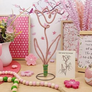 Puede incluir: Una escultura de alambre tejido de color rosa con forma de flor de tulipán, que presenta una base tejida verde y un soporte de metal circular. Junto a ella hay una tarjeta de instrucciones blanca titulada KNIT-WRAP TECHNIQUE con un dibujo.