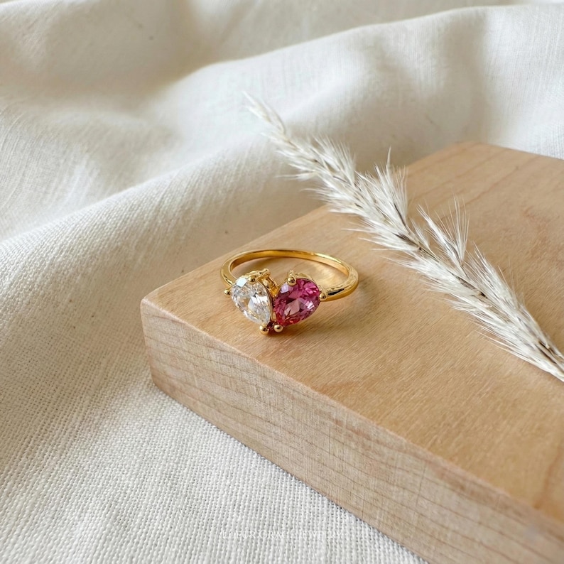 May include: A gold ring featuring two heart-shaped gemstones, one clear and one pink. The ring is set on a wooden block with a sprig of dried grass in the background. The ring's band is thin and delicate.